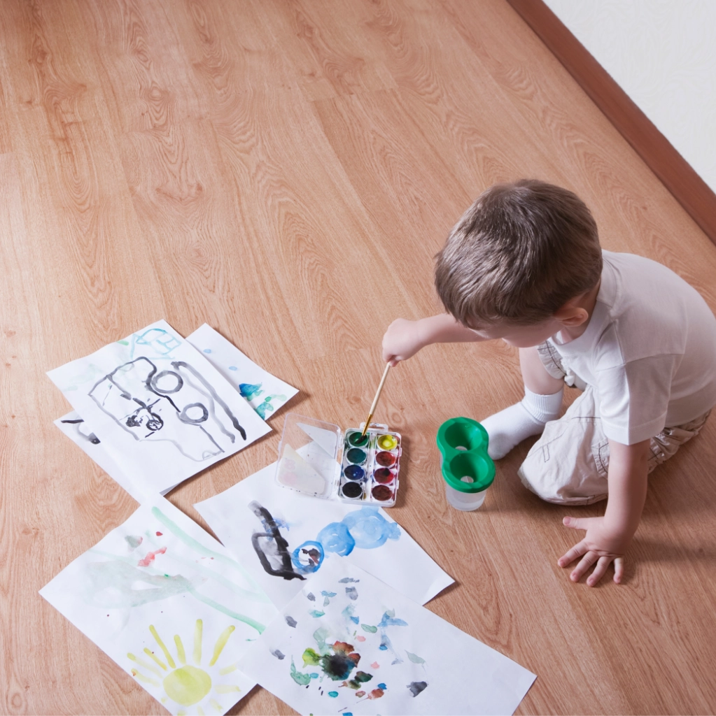 Overhead view of a young boy sitting on a wood floor painting with watercolors on several sheets of paper.